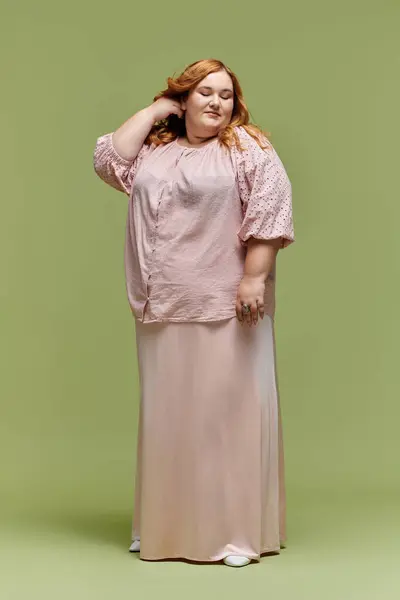 A woman in a pink top and skirt stands against a green backdrop, adjusting her hair. — Stock Photo