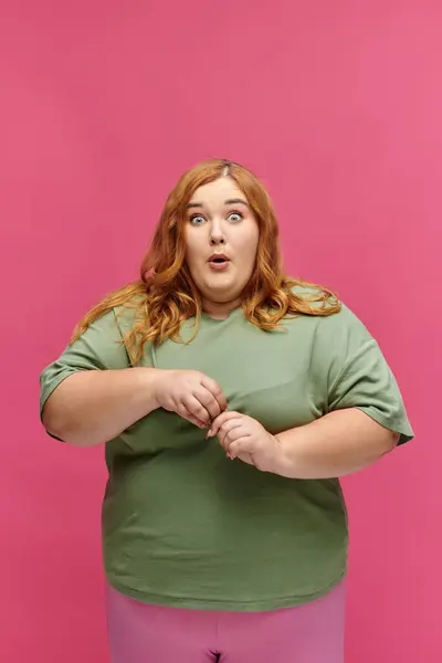 A woman in a green shirt and pink pants stares in astonishment against a vibrant pink backdrop. — Stock Photo