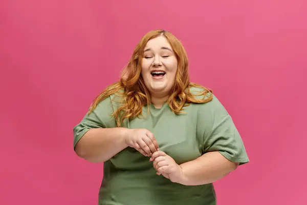 A woman in a green shirt laughs with her eyes closed against a bright pink background. — Stock Photo