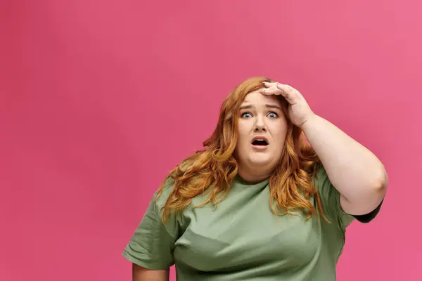 Uma mulher plus-size em uma camisa verde parece chocada com um fundo rosa. — Fotografia de Stock