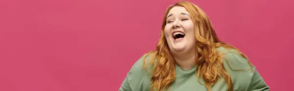 A plus size woman in a green shirt laughs heartily against a pink background. — Stock Photo