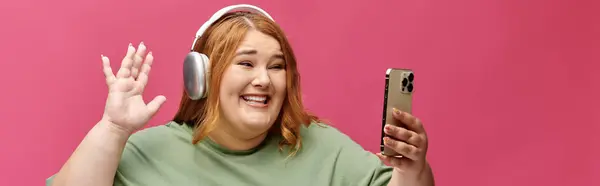 A woman with long red hair waves while smiling and looking at her phone. — Stock Photo