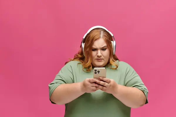 Une femme aux cheveux roux et écouteurs regarde attentivement son téléphone. — Photo de stock