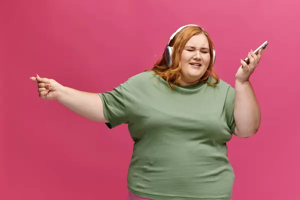 A woman in a green shirt dances with headphones on and a phone in her hand. — Stock Photo