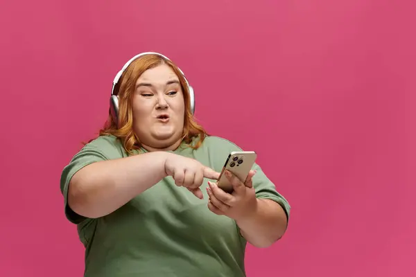 A woman wearing headphones and a green shirt uses her phone against a pink background. — Stock Photo