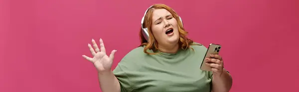 Una donna con i capelli rossi canta insieme alla musica mentre indossa le cuffie e tiene un telefono. — Foto stock