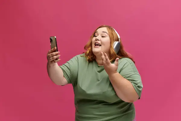 Una donna con i capelli rossi che indossa le cuffie canta entusiasta mentre tiene in mano uno smartphone. — Foto stock