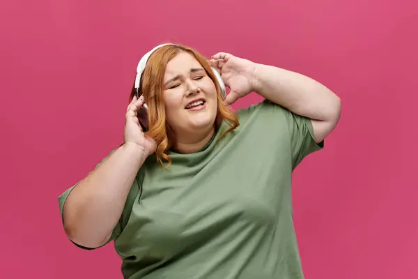 Una donna con i capelli rossi canta insieme alla musica mentre indossa cuffie e una t-shirt verde. — Foto stock