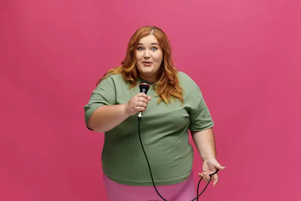 A plus size woman sings into a microphone in front of a vibrant pink backdrop. — Stock Photo