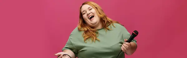 A plus-size woman laughs while holding a microphone, against a pink background. — Stock Photo