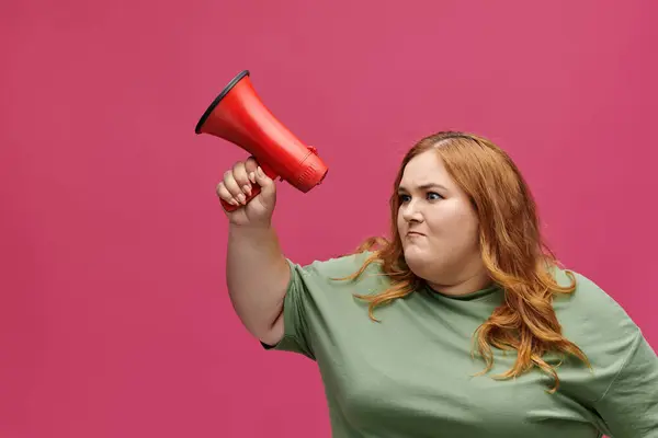 A plus size woman with red hair holds a red megaphone in front of a pink background. — Stock Photo