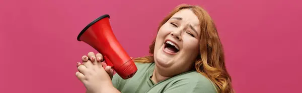 A woman with long red hair is shouting into a red megaphone with a smile on her face. — Stock Photo