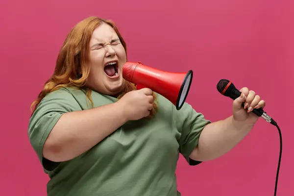 Una mujer con el pelo largo y rojo grita en un megáfono, con los ojos cerrados y la boca abierta. - foto de stock