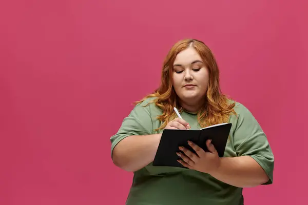 Женщина plus-size пишет в черной тетради, в зеленой рубашке на розовом фоне. — стоковое фото