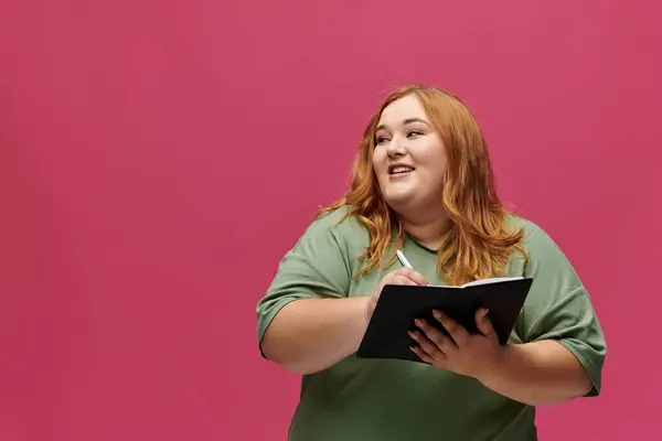 Uma mulher com uma camisa verde escreve em um caderno contra um pano de fundo rosa. — Fotografia de Stock