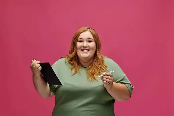 A plus size woman smiles while holding a notebook and pen in front of a pink background. — Stock Photo