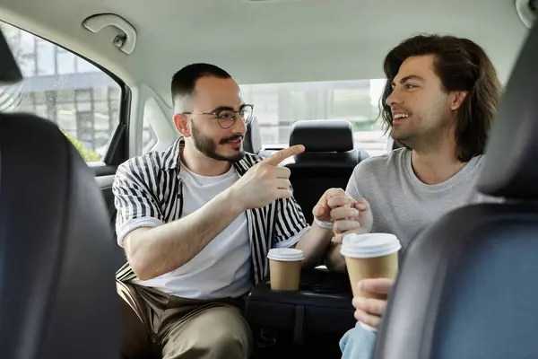 Two men, holding hands and enjoying coffee, converse in the backseat of a car. — Stock Photo