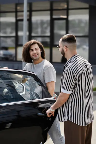 A gay couple stands beside a black car, one holding the door open. — Stock Photo