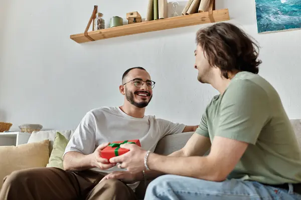 A gay couple shares a tender moment on a sofa, one presenting a gift. — Stock Photo