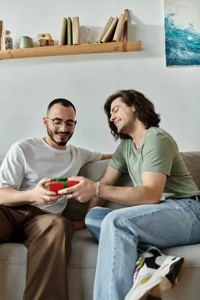 A gay couple sits on a couch, one presenting a gift to the other while both smile happily. — Stock Photo