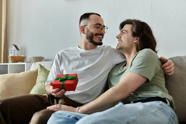 A gay couple on a couch, one man holding a wrapped gift. — Stock Photo