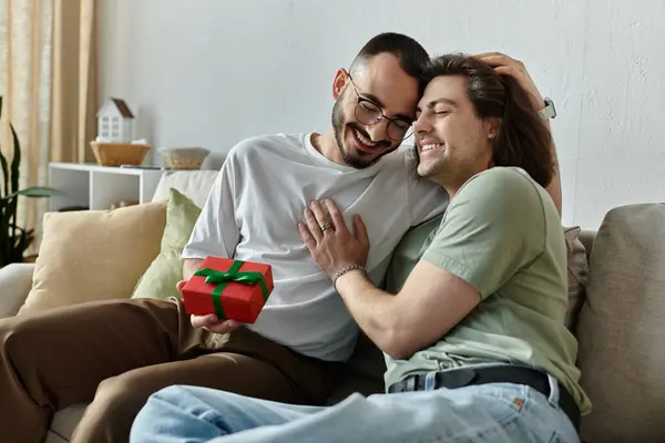 Two men cuddle on a couch, one presenting a wrapped gift to the other. — Stock Photo