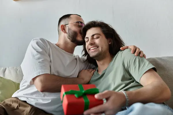 Una coppia gay si rilassa su un divano, uno con in mano un regalo mentre l'altro gli sussurra nell'orecchio. — Foto stock