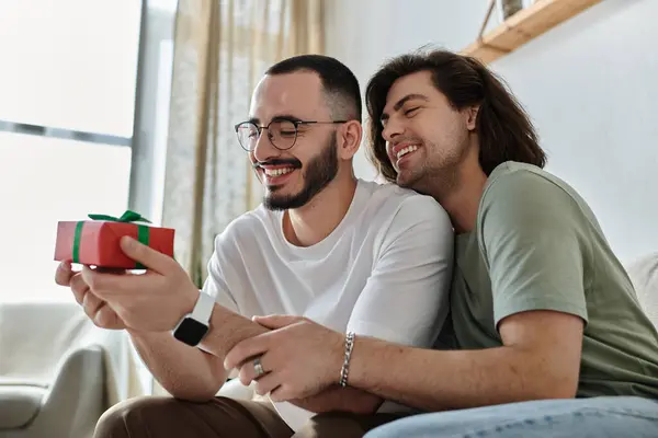 A gay couple embraces as one opens a gift, smiling brightly. — Stock Photo