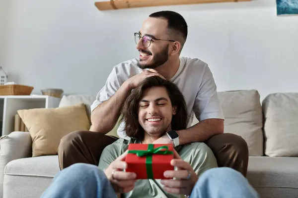 A loving gay couple sits on a couch, one man presenting a gift to the other. — Stock Photo