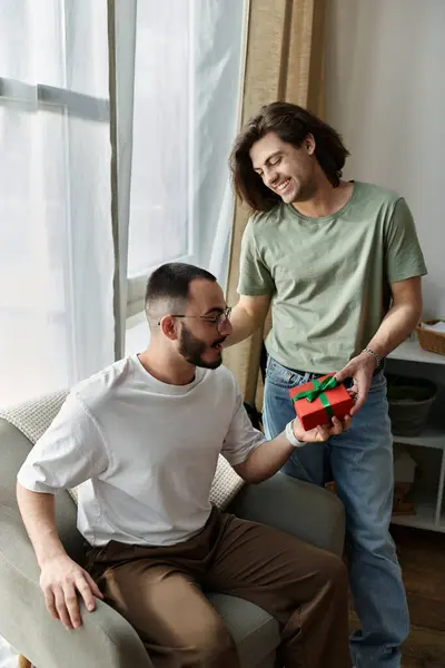 A man gives a gift to his partner, who sits on a chair, both smiling happily. — Stock Photo