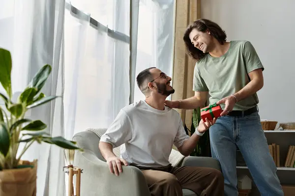 A man presents a gift to his partner, who is sitting in a chair, both looking happy and excited. — Stock Photo