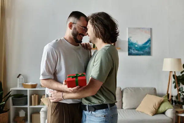 A loving gay couple embraces in a cozy living room, sharing a gift and a smile. — Stock Photo