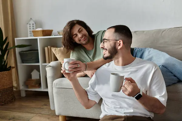 Two men sit on a couch, holding mugs of coffee and smiling at each other. — Stock Photo