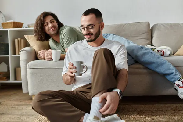 Gay couple at home, one man sits holding a mug, the other lies on the couch — Stock Photo