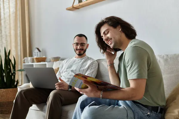 Una coppia gay si rilassa sul divano, uno lavora su un computer portatile mentre l'altro legge una rivista. — Foto stock