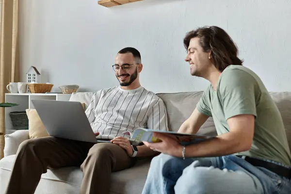 Zwei Männer entspannen sich auf einer Couch, einer arbeitet an einem Laptop, während der andere eine Zeitschrift liest. — Stockfoto