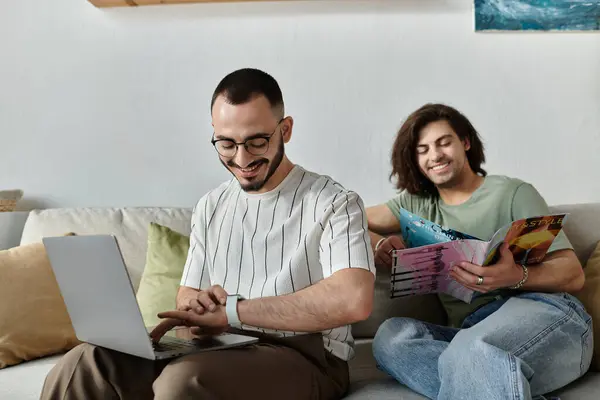 A gay couple relaxes at home, one working on a laptop, the other reading a magazine. — Stock Photo
