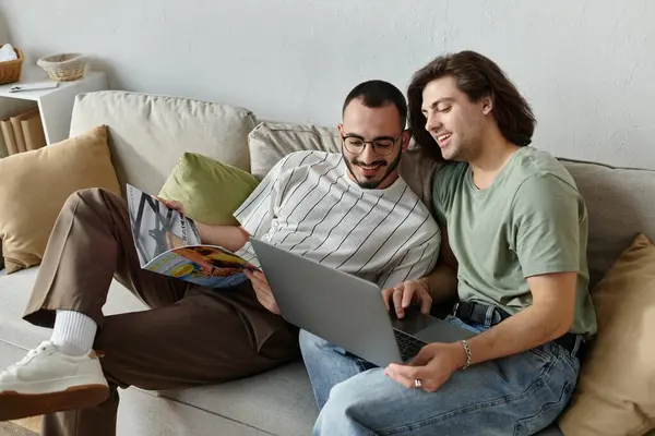 A loving gay couple relaxes on a couch, one reading a magazine and the other using a laptop. — Stock Photo