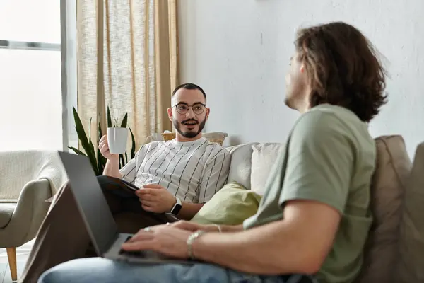 Dois homens em um sofá, um com café, o outro em um laptop, desfrutando de companhia — Fotografia de Stock