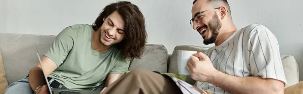 A gay couple on a couch, one looking at a laptop while the other laughs and holds a mug. — Stock Photo
