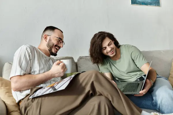 Two men laugh together while sitting on a couch, one holding a cup and the other using a laptop. — Stock Photo