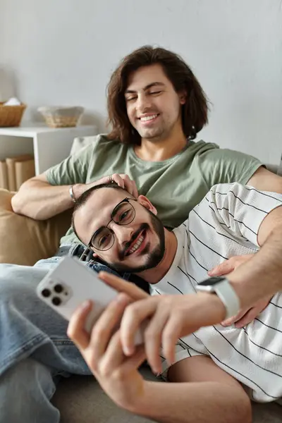 A gay couple relaxes on a couch, one laying with his head in the other's lap as they use a phone. — Stock Photo