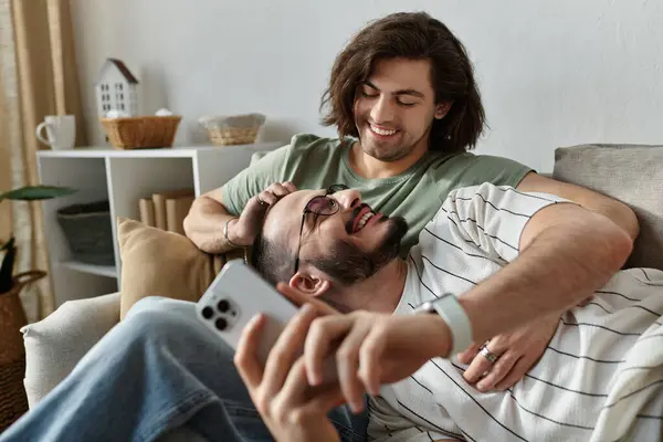A couple laughs together on a couch, one man with his arm around the other. — Stock Photo