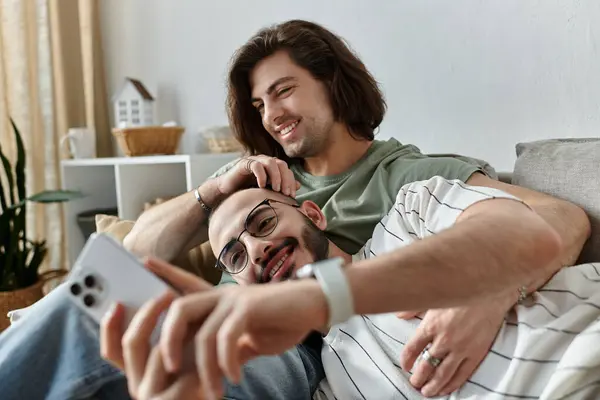 A gay couple cuddles on a couch, one man looking at his phone while the other smiles at him. — Stock Photo