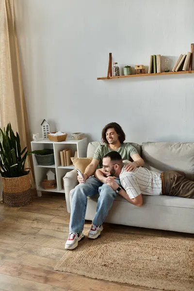 Two men relax on a couch, one lying down while the other sits and looks at a phone. — Stock Photo