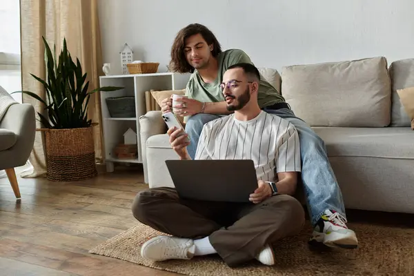 Two men relax on the floor, one using a laptop while the other leans against a sofa and holds a mug. — Stock Photo