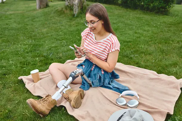 Eine junge Frau mit Beinprothese entspannt sich auf einer Decke in einem Park und scrollt auf ihrem Handy. — Stockfoto