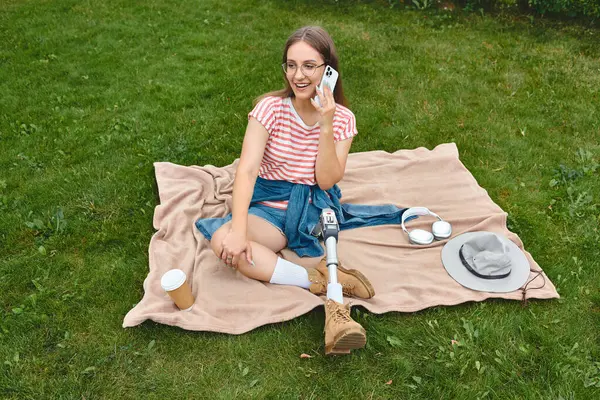 A young woman with a prosthetic leg sits on a blanket in a park, smiling and talking on her phone. — Stock Photo