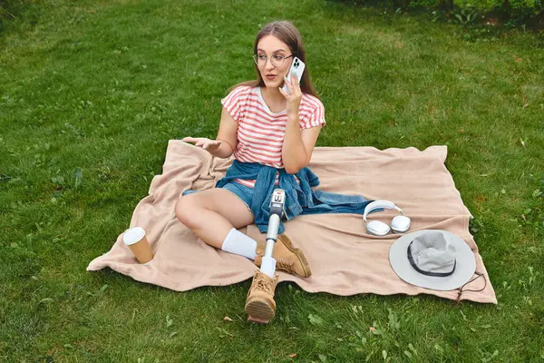A young woman with a prosthetic leg sits on a blanket in a park, talking on her phone. — Stock Photo