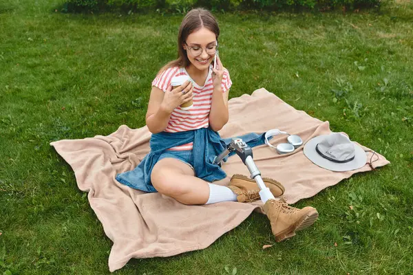 A young woman with a prosthetic leg enjoys a summer day, sitting on a blanket in a park while talking on the phone and enjoying a drink. — Stock Photo